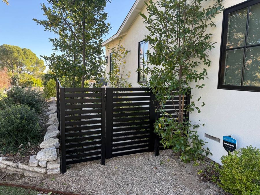 Black slatted gate beside a stucco house along a gravel path with stone edging and trees
